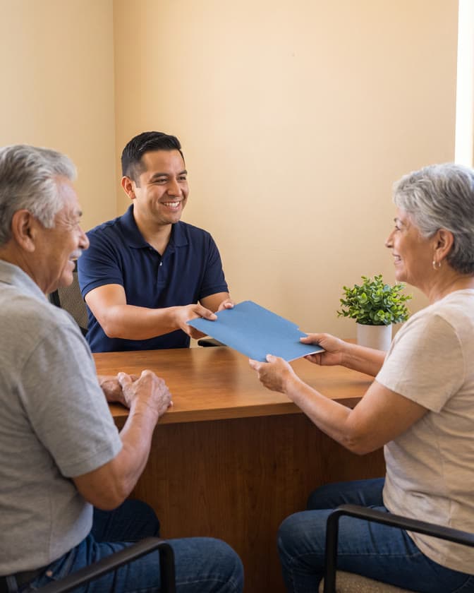 A social services staff member hands a blue folder across a desk to an older couple